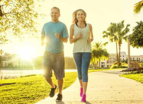 A man and a woman jogging along a paved path in a sunny park with palm trees and a lake in the background.